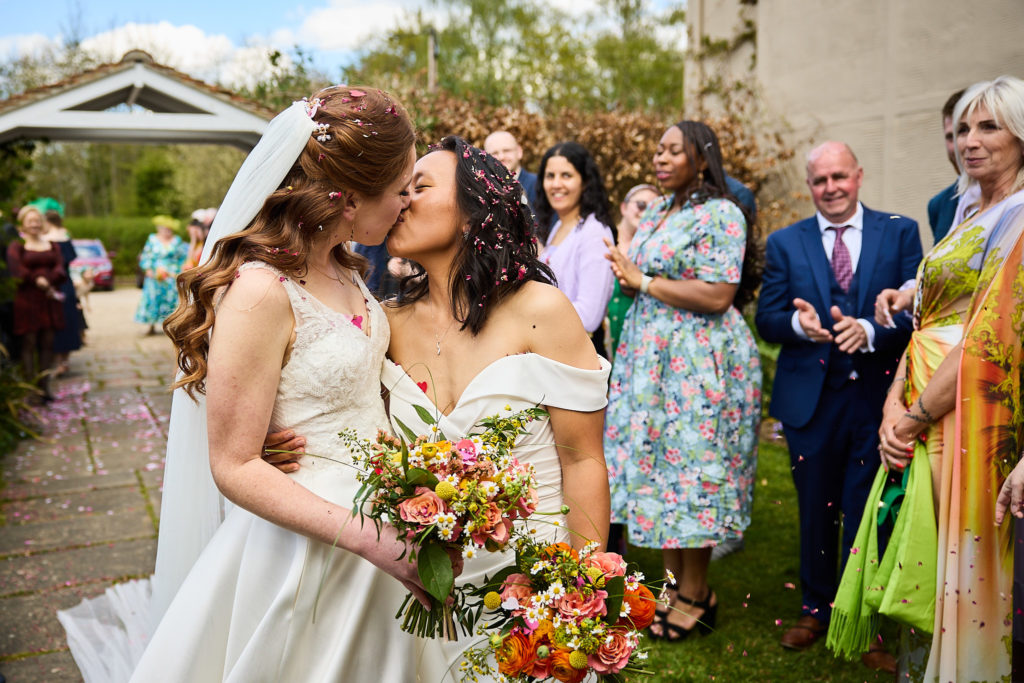 Two brides kissing as guests in colourful outfits look on at their gay wedding in Essex