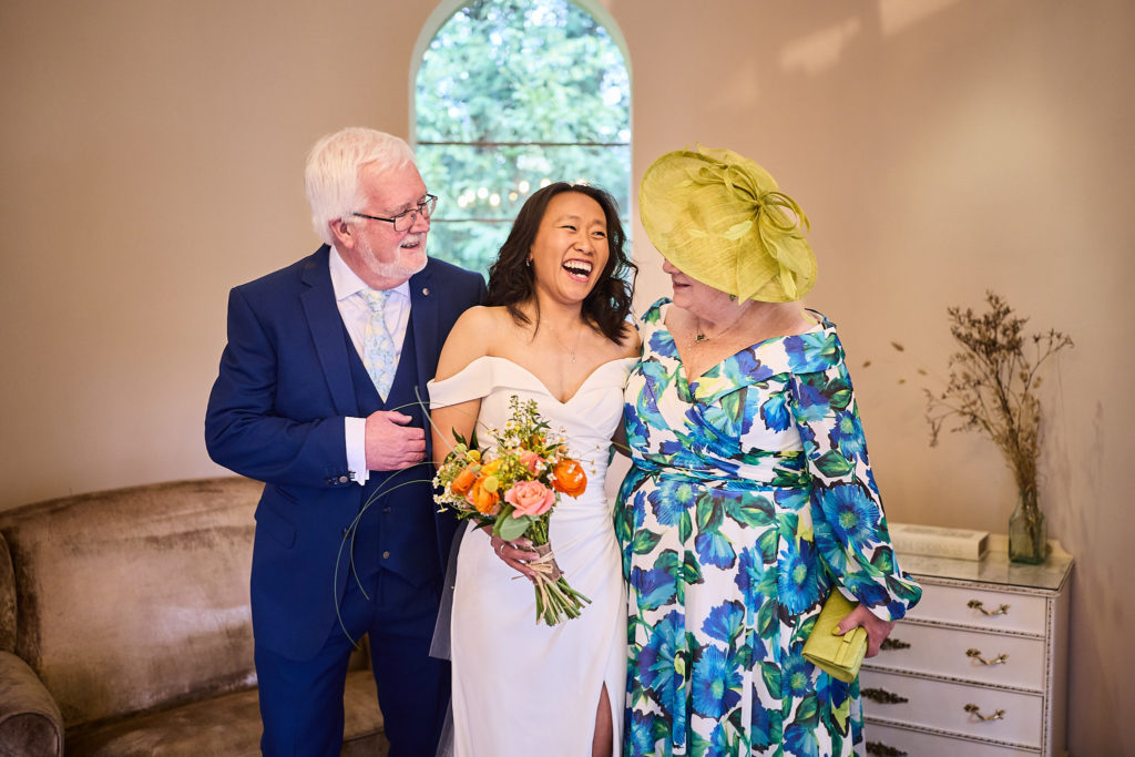 Bride laughing between an older man in a navy suit and a woman in a floral dress and yellow hat