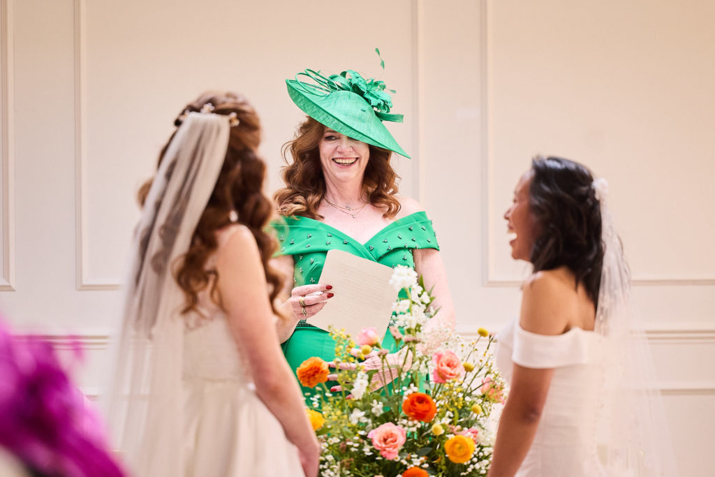 Two brides laughing with a guest in a green dress and matching hat