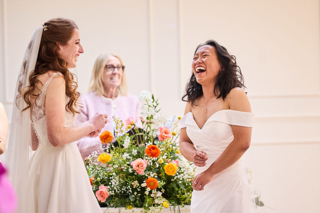 Two brides laughing with the registrar behind a display of flowers