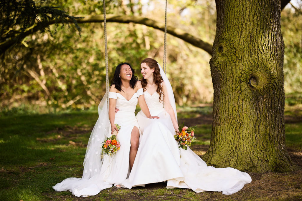 Two brides sitting together on a tree swing at their same-sex wedding at Prested Hall