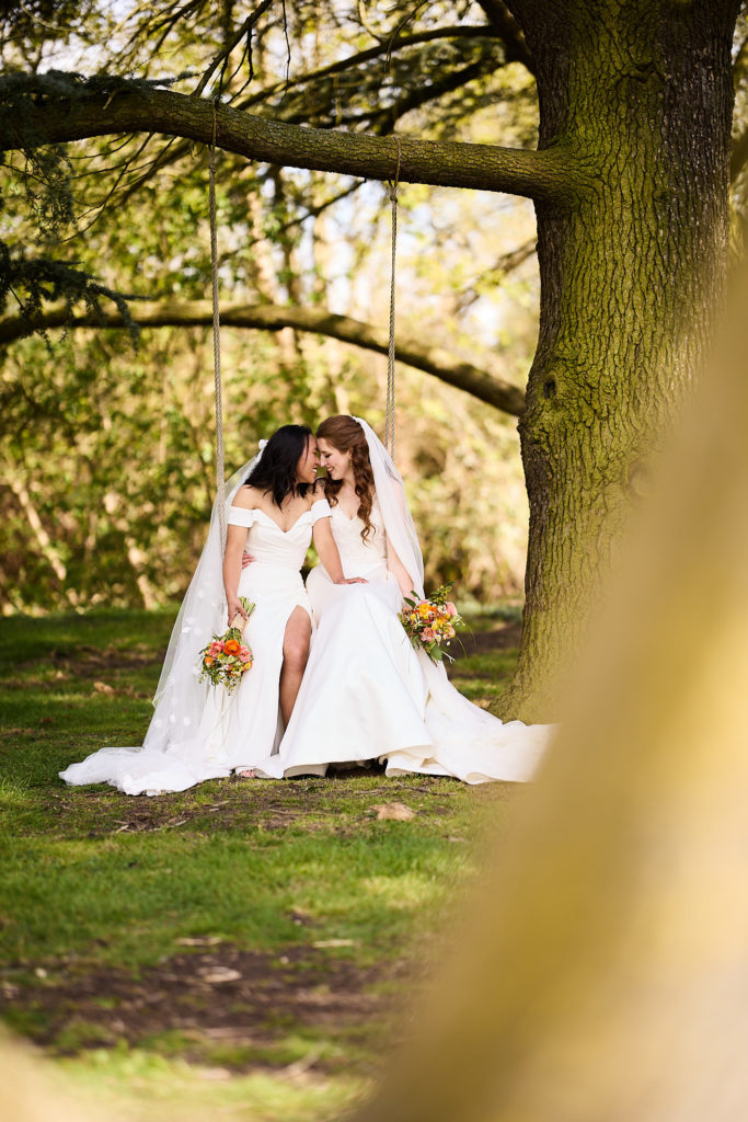 Two brides kissing on a tree swing in the grounds of Prested Hall