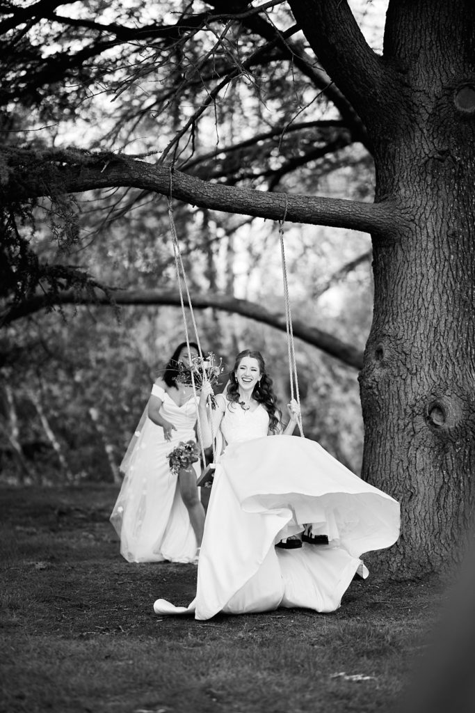 Black and white photo of a bride swinging on a tree swing as her wife stands beside her