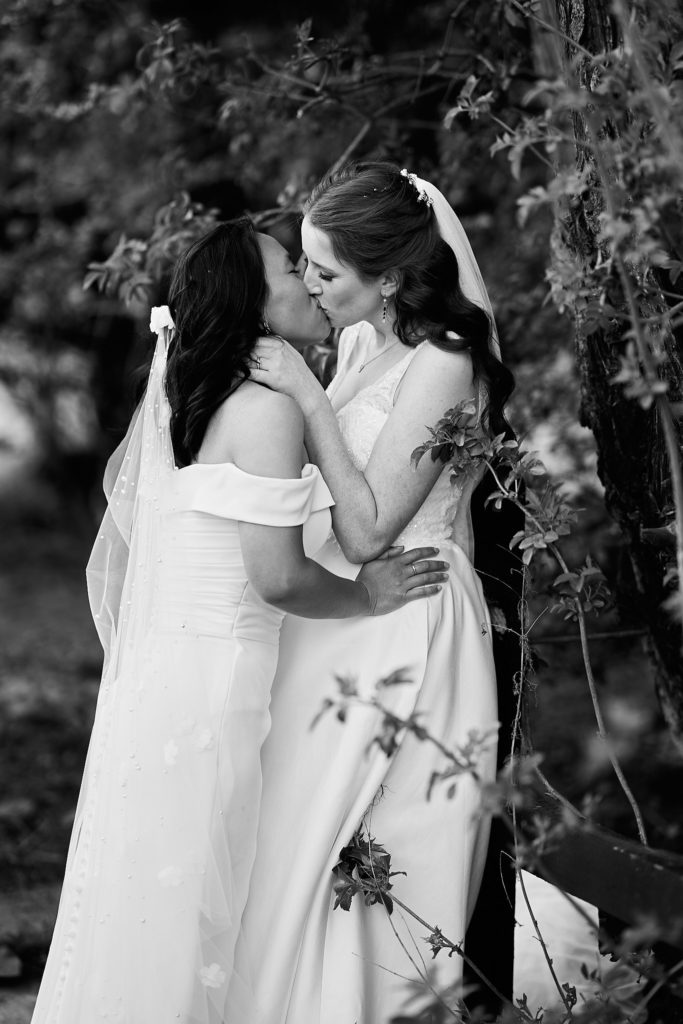 Black and white photo of the two brides kissing among the trees at Prested Hall