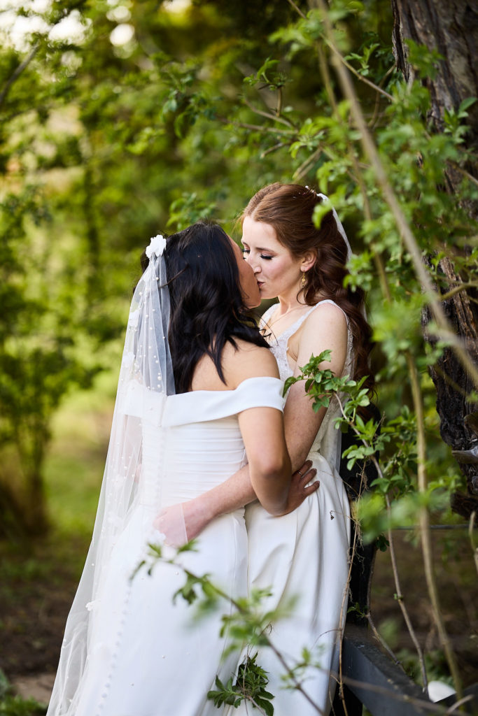 Two brides kissing among spring leaves at Prested Hall