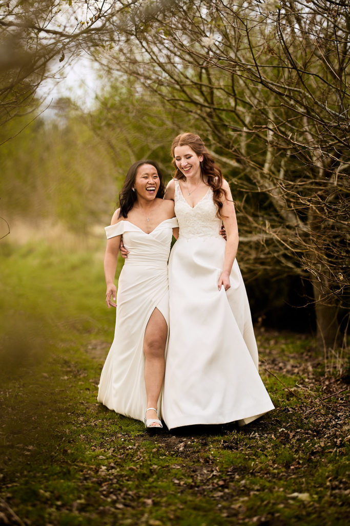Two brides walking arm in arm along a woodland path at Prested Hall