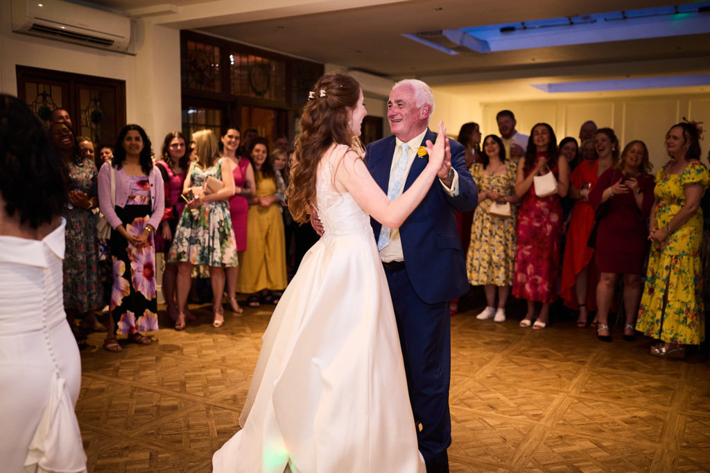 Bride dancing with an older man in a navy suit as guests watch