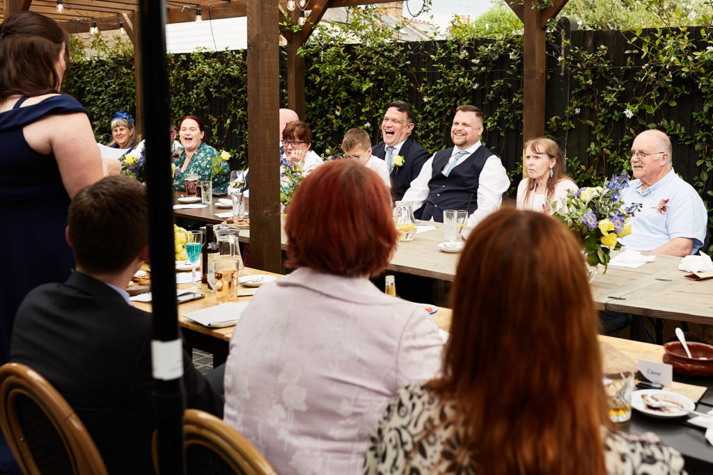 Wedding guests seated at long tables under a wooden pergola for the wedding breakfast