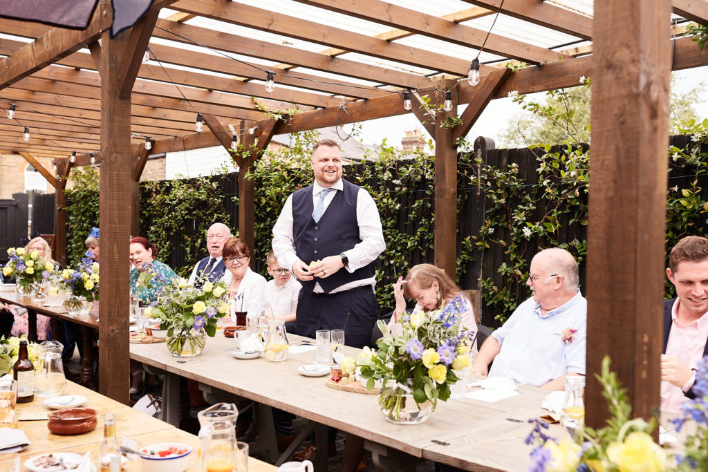 Groom in a waistcoat giving his speech to guests under a pergola