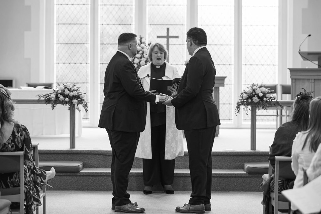Black and white photo of two grooms holding hands in front of the minister during their wedding ceremony