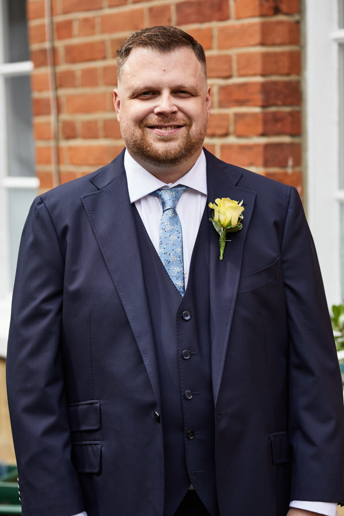 Groom smiling in a navy three-piece suit with a pale blue tie and yellow rose buttonhole