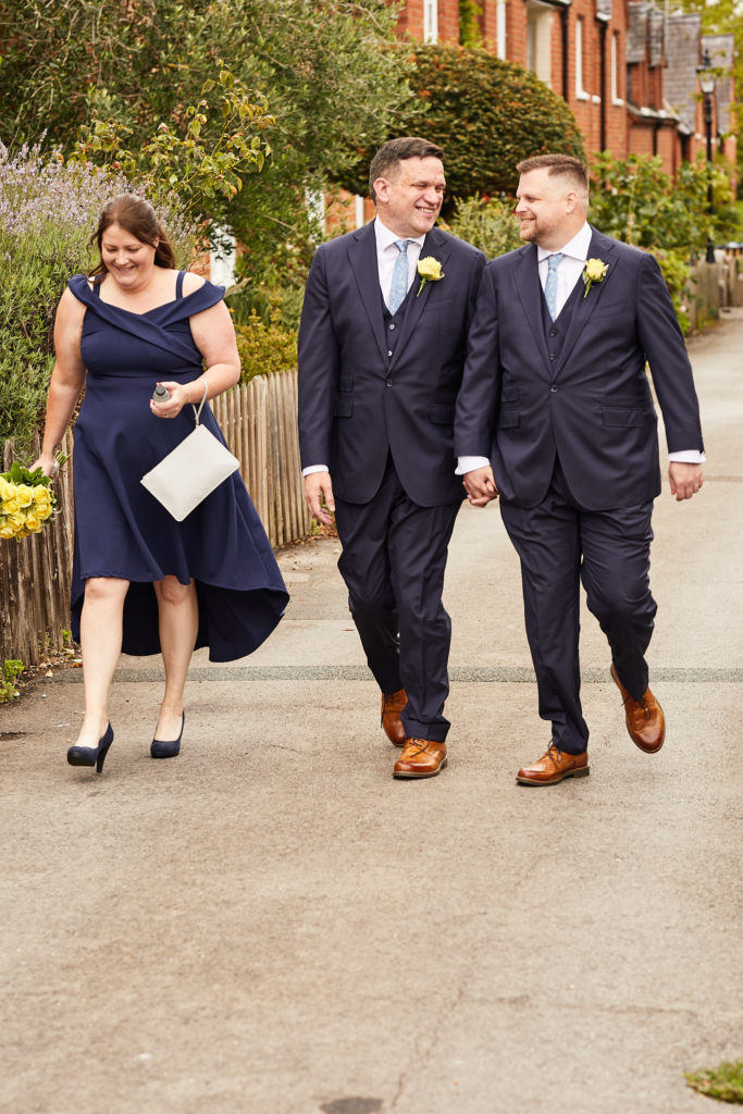 Two grooms walking hand in hand along the street with their best woman beside them