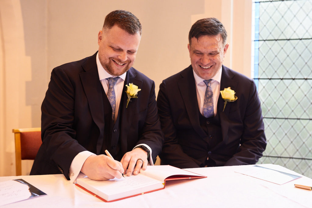 Two grooms smiling as they sign the register