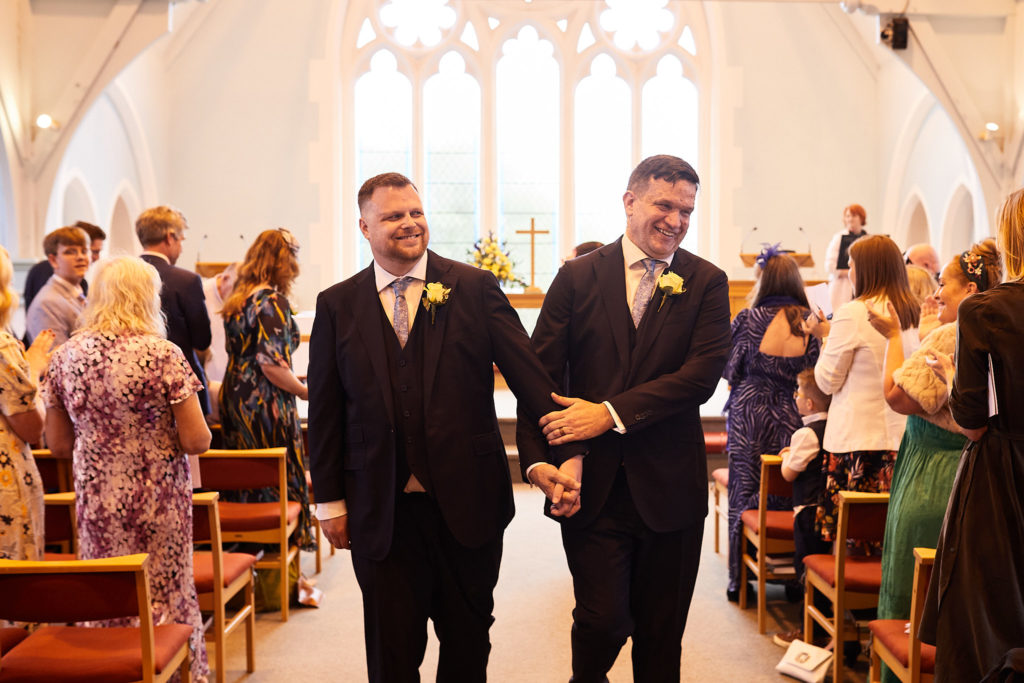 Two grooms walking back down the aisle together as guests applaud at their same-sex wedding