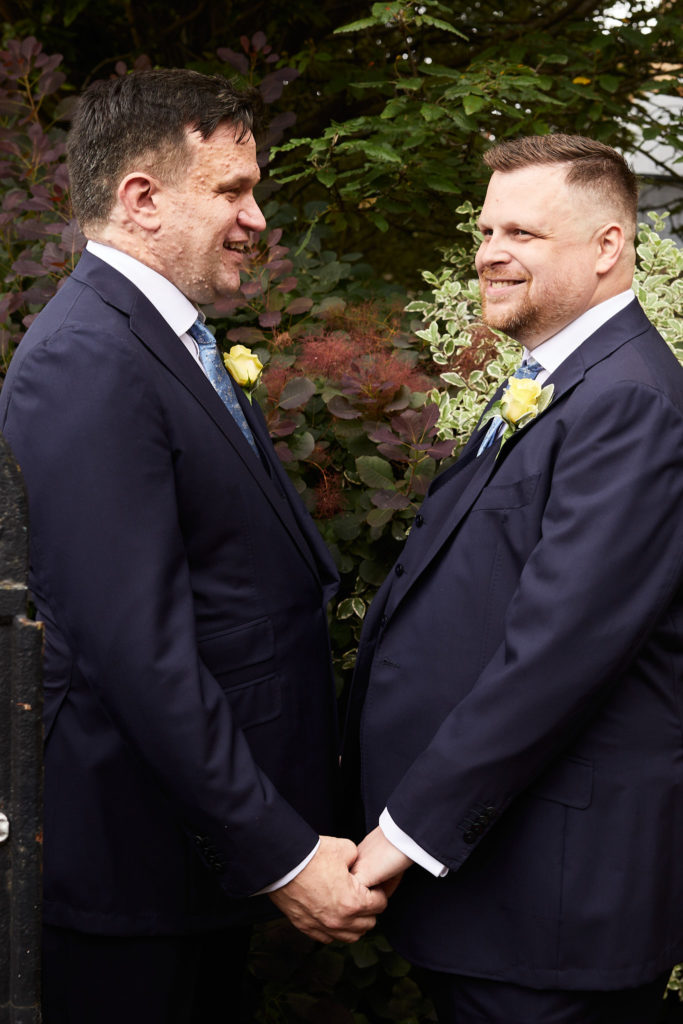 Two grooms holding hands and smiling at each other on their gay wedding day