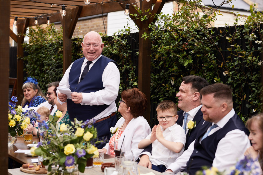 Guest in a waistcoat giving a speech as the grooms listen at the table