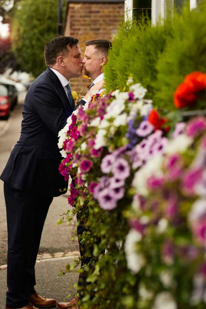 Two grooms about to kiss beside hanging baskets of flowers