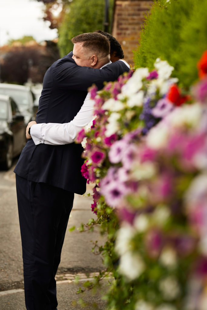 Two grooms hugging beside baskets of pink and white flowers