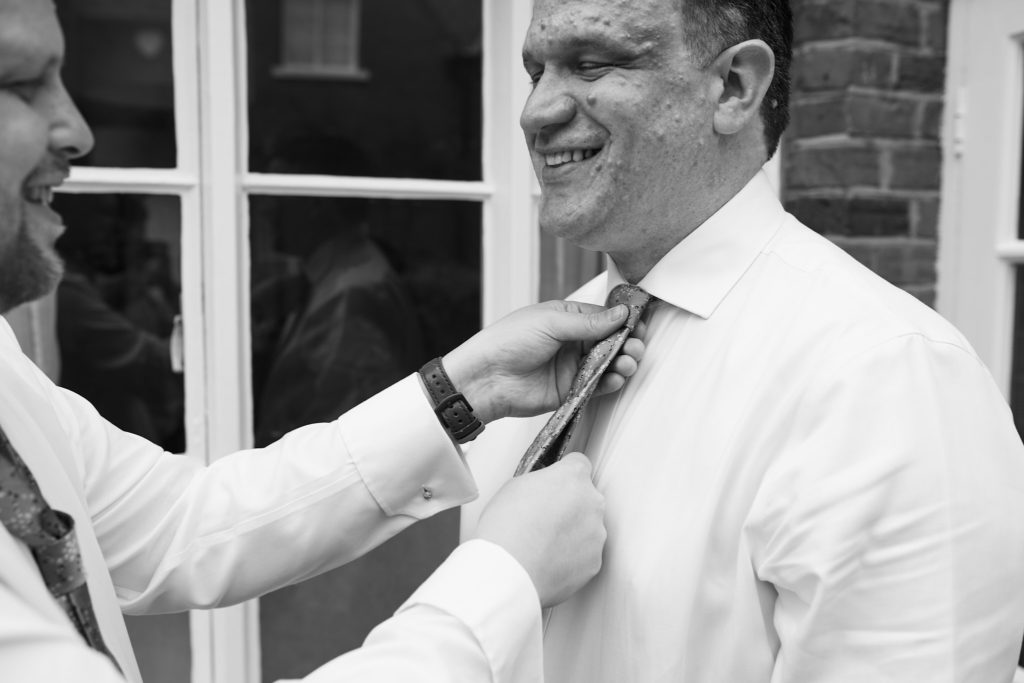 Black and white photo of a groom laughing as his tie is straightened