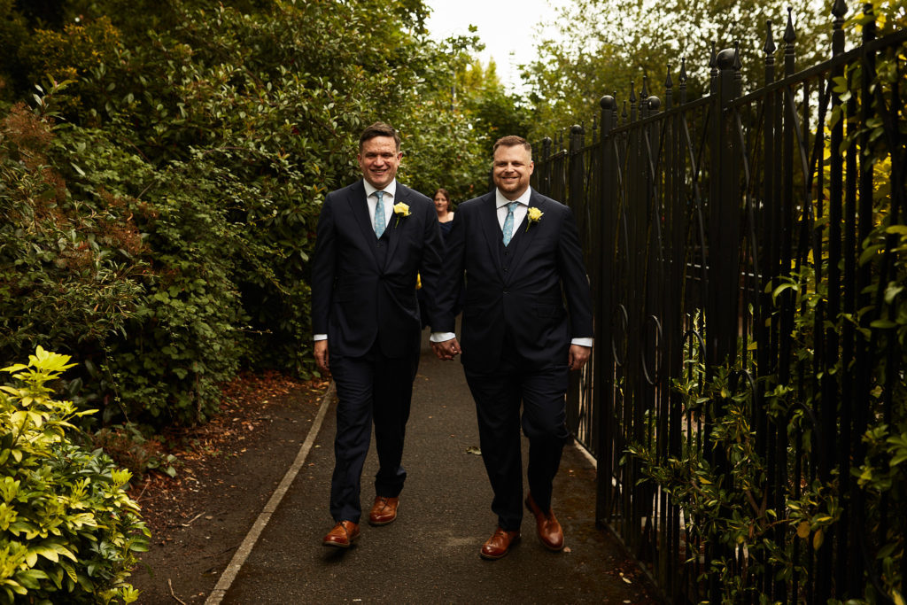 Two grooms walking hand in hand along a tree-lined path