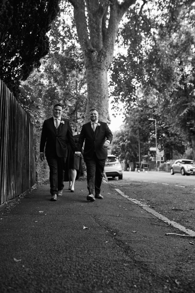 Black and white photo of the two grooms walking hand in hand along a tree-lined road