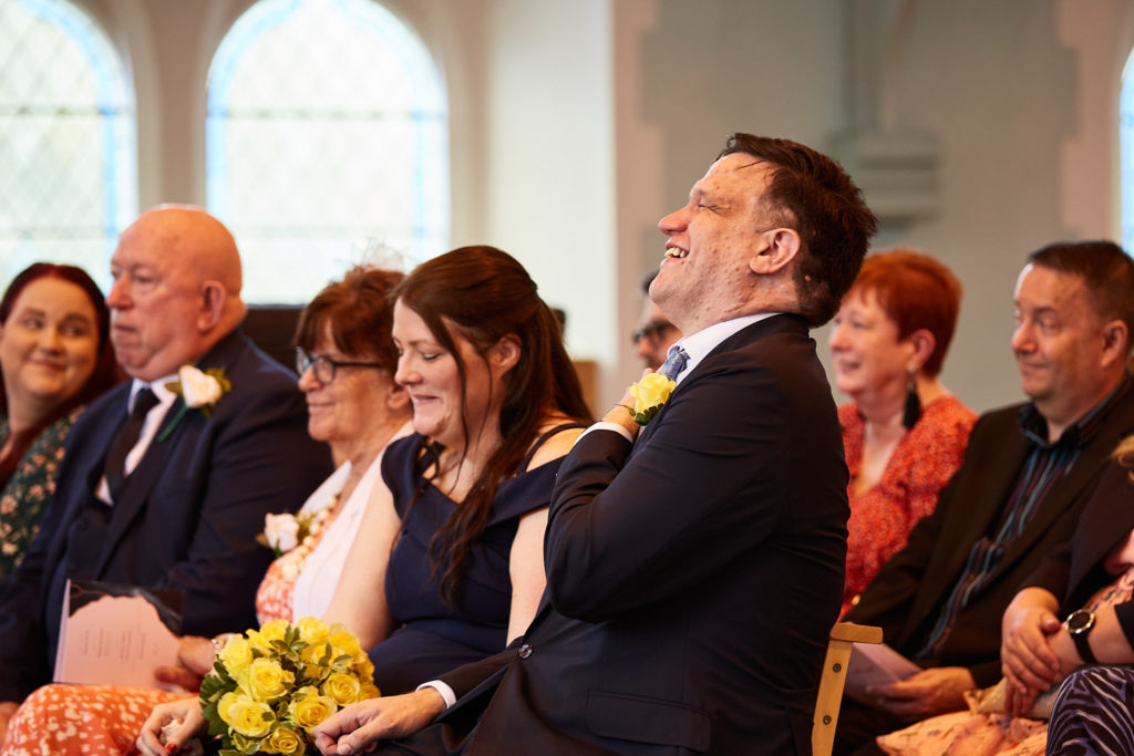 Groom laughing as he sits among guests during the ceremony