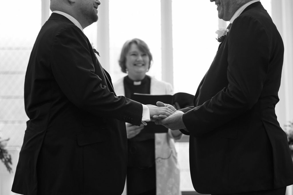 Black and white photo of the two grooms exchanging rings in front of the minister