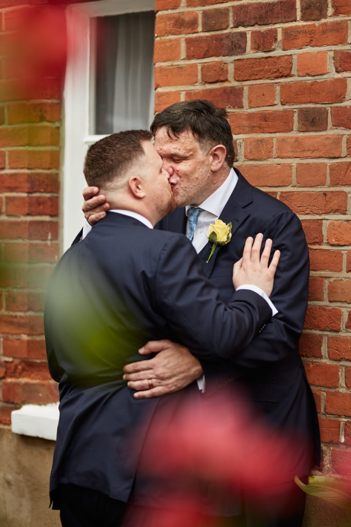 Two grooms kissing against a red-brick wall, seen through flowers