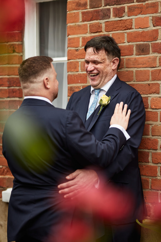 Groom laughing against a red-brick wall with his husband's hand on his chest