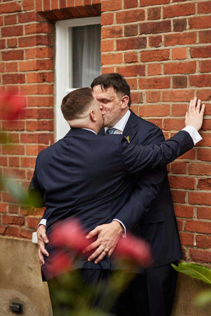 Two grooms kissing against a red-brick wall with red flowers in the foreground
