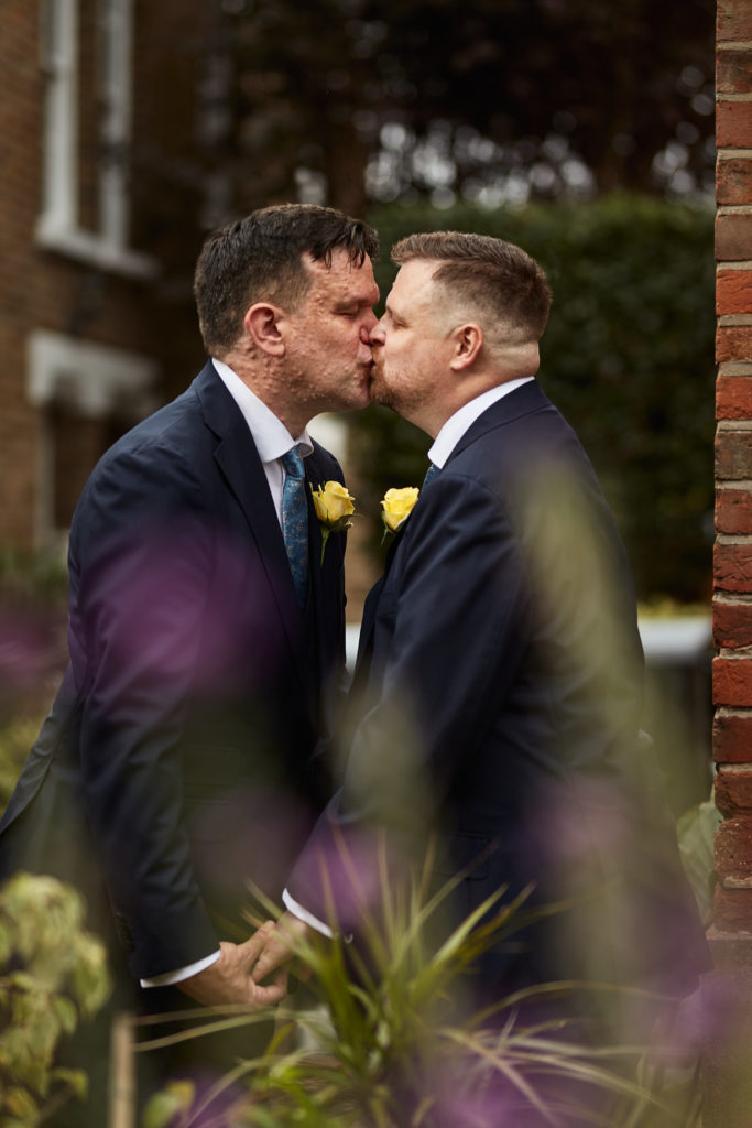 Two grooms kissing among garden flowers on their same-sex wedding day