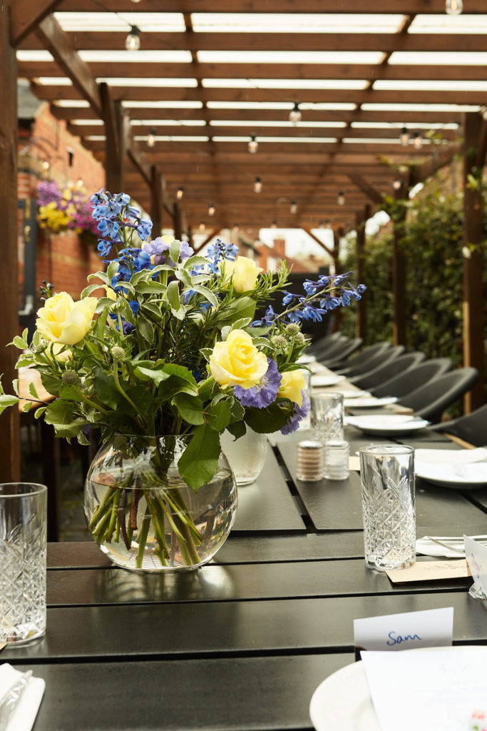 Long table set for the wedding breakfast under a pergola, with yellow roses and blue flowers