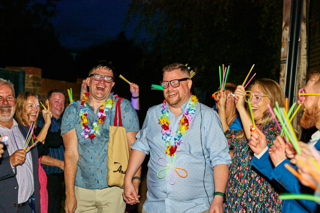 Two grooms in matching shirts and flower garlands walking past guests waving glow sticks