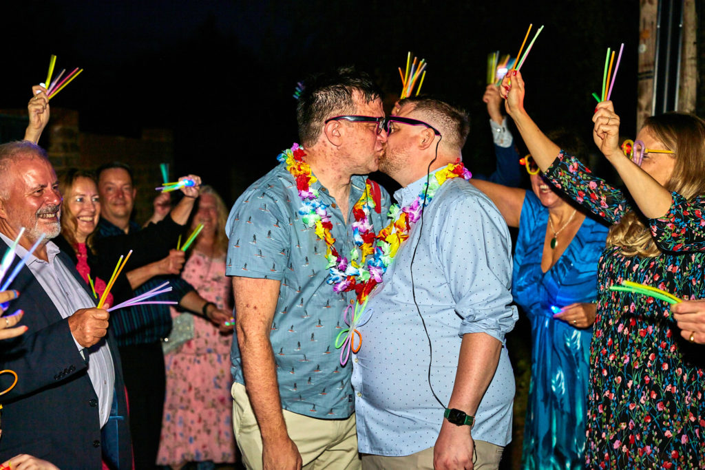 Two grooms kissing in matching shirts and flower garlands as guests cheer with glow sticks