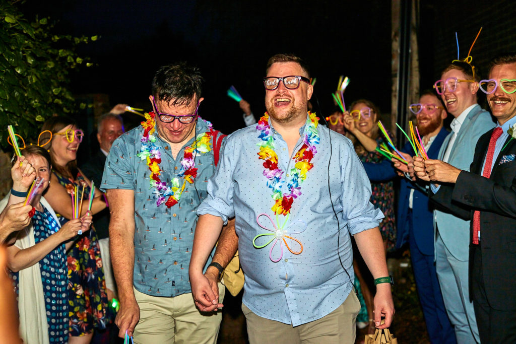 Two grooms laughing in flower garlands as guests wave glow sticks at their LGBT wedding party