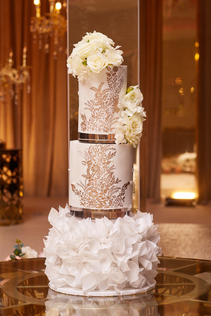 Tall white wedding cake with gold lace detail, ruffled base and white flowers