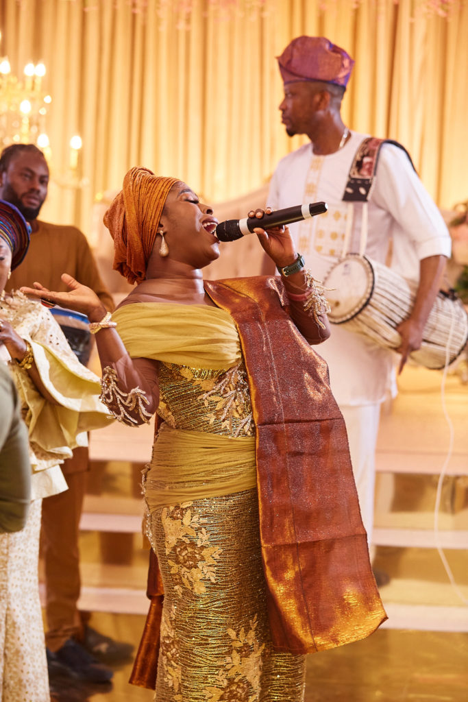 Singer in a gold head tie performing with a talking drum player at a traditional wedding