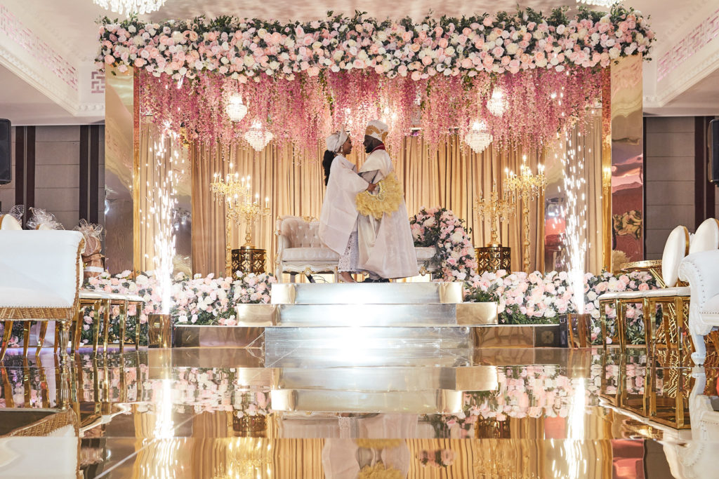 Bride and groom on the wedding stage beneath a canopy of flowers, seen across the mirrored floor