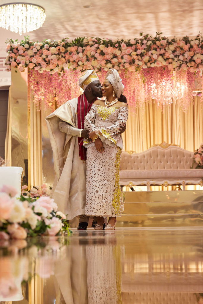 Groom embracing the bride on stage in front of the flower wall and gold drapes