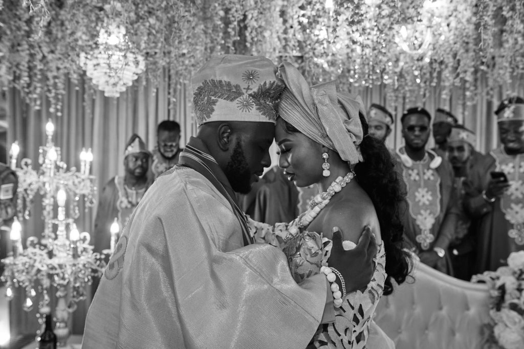Black and white photo of the bride and groom forehead to forehead beneath chandeliers