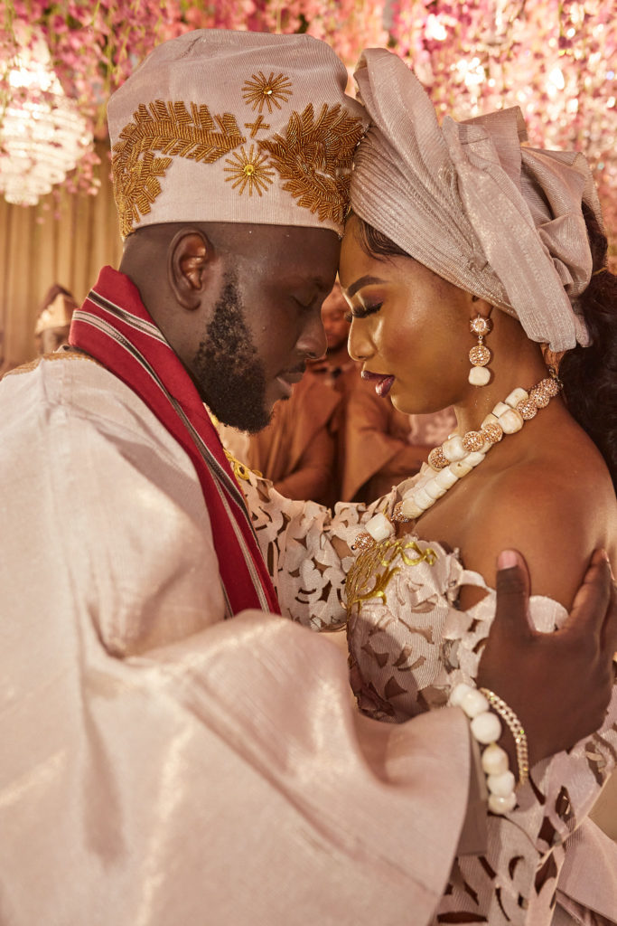 Bride and groom forehead to forehead in cream traditional attire and coral beads