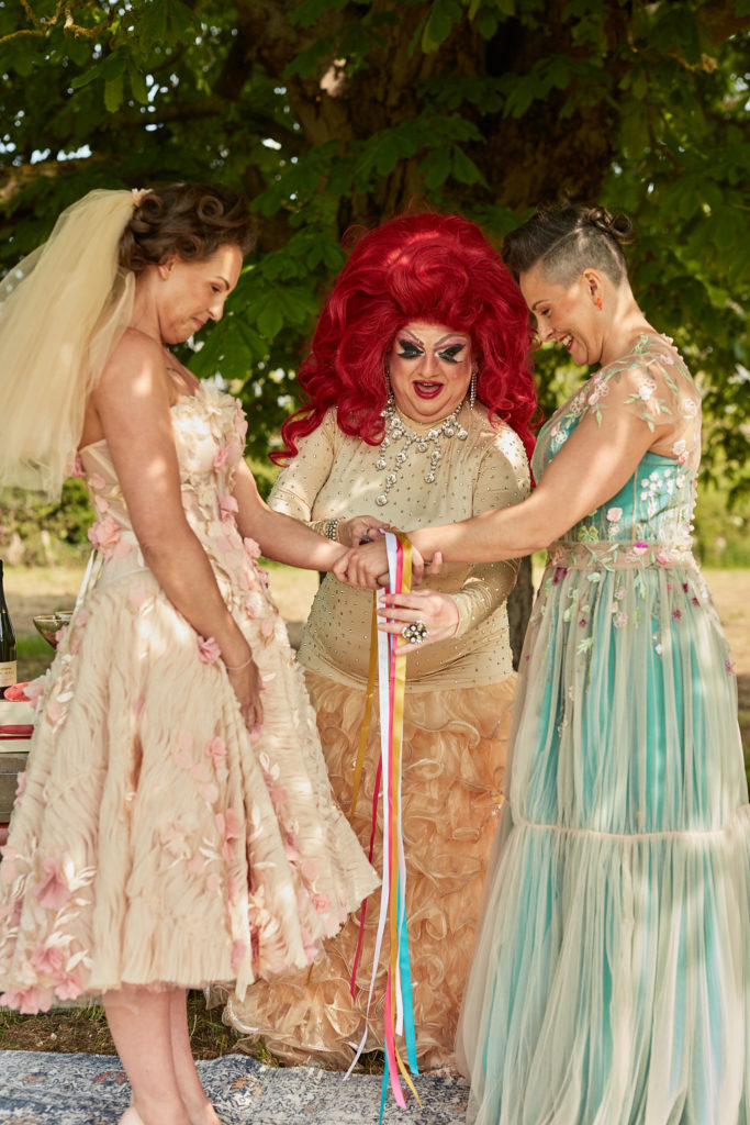 Two brides having their hands bound with ribbons by a drag queen celebrant