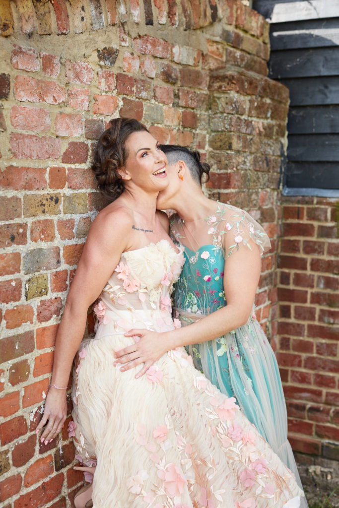 Bride kissing her laughing wife's neck against a red-brick wall