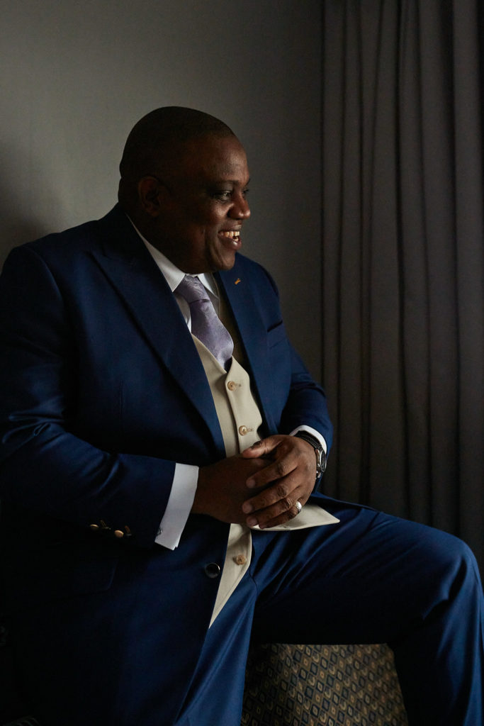 Groom in a navy suit smiling as he sits by a window before the wedding