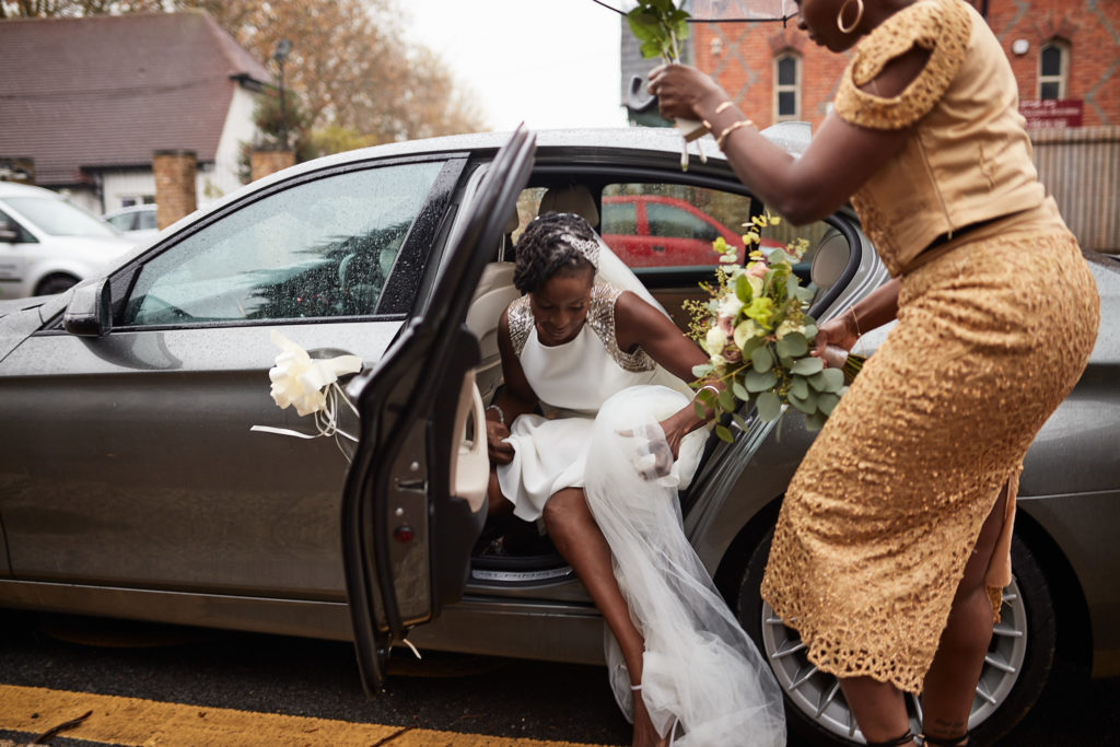 Bride being helped out of the wedding car by a woman in a gold lace dress