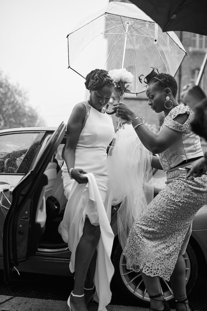 Black and white photo of the bride arriving under an umbrella as her dress is adjusted